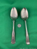 Four silver antique serving spoons arranged in pairs on green background with quarter coin for scale showing front side with ornate and plain handles
