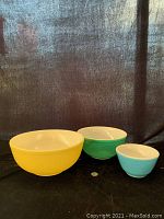 Three nested vintage Pyrex mixing bowls in yellow, green, and blue with white interiors shown side by side on black cloth background, coin for scale.