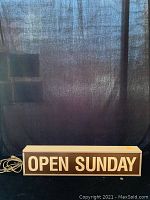 Front view of rectangular gray sign with white letters reading OPEN SUNDAY
