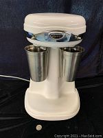 Front view of white Hamilton Beach milkshake machine with two attached stainless steel cups and chrome band with badge.