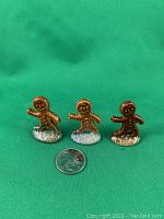 Three small gingerbread men figurines on white bases, with quarter coin for size reference, front view