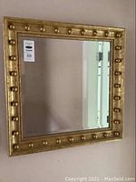 Full view of the square mirror hanging on the wall showing the gold frame with round studs.