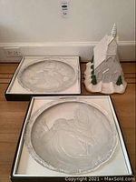Two Mikasa ceramic Christmas plates in original boxes with stands and a white ceramic church decoration on the floor.