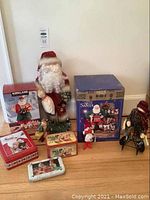 Full view of Christmas Decor lot showing multiple Santa statuettes, tins, playing cards and a figurine of a person in dark outfit.