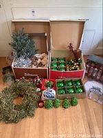 Full lot overview showing boxes of green and gold baubles, the wreath, glass candle votives, and assorted decor items including small green pine cones.