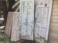 Five vintage wood doors with peeling white paint, showing worn condition. Some doors include hardware such as hooks and knobs. The doors have rectangular panels.