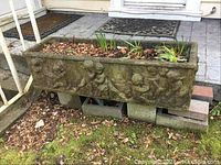 Front and side views of the rectangular cement planter with cherub relief showing the entire item and its placement on cement blocks.