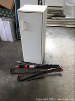 White metal storage cabinet with handle. Crown bars, Shur-Line extension handle, and golf swing practice device placed in front of cabinet on concrete floor.