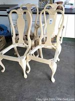 Four beige wooden chair frames in a group, two with arms and two without arms, showing curved backs, carved shell detail at top, and transparent seat bases.