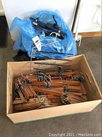 Box containing many wooden hangers with metal clips and several wire pant racks stacked inside a cardboard box on the floor.