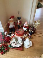 Photo showing full lot of Christmas decor including figurines, plates, and containers arranged on wood floor near white wall corner.
