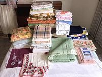 Multiple folded vintage textiles including tablecloths, sheets, wool blanket, and curtains stacked on a bed