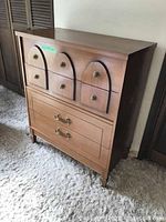 Side angle view of wooden mid century style dresser, showing front and left side, featuring three top drawers with curved fronts and metal ring pulls, larger bottom drawer with metal handles, on carpeted floor.