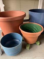 Four pots on floor: large terracotta, medium terracotta with green interior on wooden stand, small light blue plastic, large navy-blue plastic