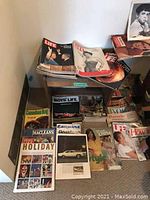 Overview of vintage magazines stacked on and beside a wooden side table and wooden bench against a wall. Visible titles include Life, Boys Life, Vogue, Macleans, Esquire, Gossips, and others.