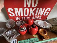 Four red apple-shaped vintage lighters and various glass and ceramic ashtrays grouped on a wooden surface under a 'No Smoking' sign. Includes a square glass ashtray and small wooden ashtray.