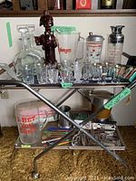 Full view of vintage bar cart showing glass decanters, shot glasses, cocktail shakers, and assorted bar tools on wooden shelves within silver metal frame with wheels.