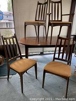 Photo showing four wooden chairs and wood dining table. Chair cushions covered in light brown upholstery. One chair is captain chair with armrests.