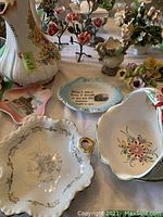 Photo showing multiple porcelain items including vases, ashtrays, and floral porcelain arrangements on a table.