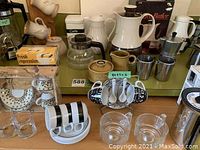 Photo showing an assortment of coffee and kitchenware items including a white Coffee Butler percolator, clear glass coffee pot, brown and beige ceramic sugar pots, metal creamers, and black and white espresso cups on a rotating stand with spoons.