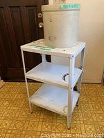 Full view of white enamel three-shelf metal kitchen cart with Medalta crock on top, placed indoors on vinyl floor.