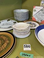 Stack of assorted plates and bowls on green tabletop, some with decorative rims and anniversary markings