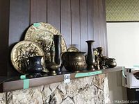 Wide angle showing assortment of brass tableware and decorative items on a mantle shelf, including large imprinted plates, candle holders, vases, and pots.