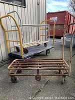 Set of 3 metal dollies, showing one rusted and two larger with shelving and yellow frames outdoors.