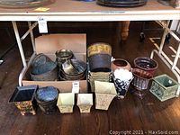 Photo taken showing the full assortment of planters and cache pots arranged on the floor under a table. Various shapes, materials, and colors shown.