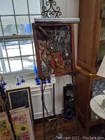 Frontal view of the large mixed media industrial art piece on decorative wrought iron easel stand with window background.