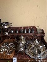 Full view of tea service, pewter fruit, silver plate loving cup, and flatware basket on a wooden shelf with beige wall background