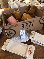 Box with mustard yellow Roscher stoneware mugs, pink textured mugs, ceramic ramekins, and white sushi trays with decorative figures.