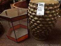View of ceramic stool and hexagonal glass stand together on carpeted floor. The ceramic stool has a distinctive bumpy surface texture, the glass stand has metal framing and reflective glass panels with one missing pane.
