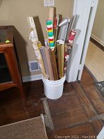 Full view of bucket filled with multiple rolls of wrapping paper and cellophane held upright.