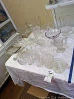 Overview photo showing various glasswares including cake stands, trifle dish, pitchers, vases, and snifters arranged on a white cloth-covered table.