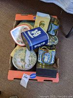 Top-down photo of multiple vintage decorative tins arranged in a box, showing various designs and sizes.