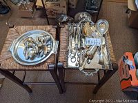 Overall lot of large silver plated serving utensils and two trays shown on two chairs, including spoons, forks, cake servers, tarnished condition.