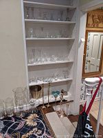 Shelf displaying various clear glass candlesticks, votive holders, small vases, and paperweights alongside brass candleholders and a metal bin