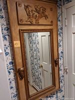 Full view of the ornate mirror showing the chariot relief at the top and mascaron candle holders on each side, alongside a door and floral wallpaper.