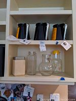 Three black coffee carafes with lids and handles on top shelf. Below shelf holds a metal kitchen container, ribbed glass container, and two glass juice jugs, one with yellow lid.