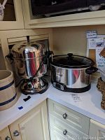 Corner kitchen countertop with large stainless steel party coffee urn with broken faucet piece removed and placed in front. Adjacent electric crock pot with stainless steel and black finish, glass top with black handle.