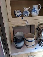 Shelf with three porcelain blue and white pitchers on upper shelf and a metal blue pitcher and ceramic crock on lower shelf.