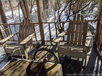 Two weathered wooden Adirondack chairs on porch with armrests and slatted backs, snow and trees in background showing outdoor setting.