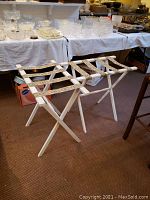 Two folding luggage racks with white wooden frames and printed canvas straps standing unfolded on carpeted floor.