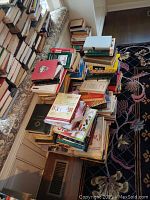 Photo showing a large assortment of books piled on a bench and stacked in various orientations. Varied covers and sizes, arranged in an unorganized pile.