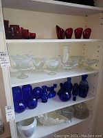 Shelf with red glass vases on upper shelf, middle shelf with opalescent and milk glass candy dishes, vases, and a tall white milk glass vase, lower shelf filled with cobalt blue glass vases and bottles as well as clear and milk glass items.