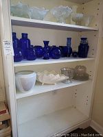 Two shelves with assorted cobalt blue glassware, iridescent bowls and clear glass dishes, white decorative bowls. A mix of vases, jars, candle holders.