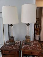 Front view of two vintage brass and glass table lamps with white drum shades standing on wooden tables.