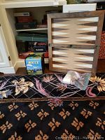 Wooden backgammon board and unopened boxed bocce ball game set on carpet with bag of game pieces in front. Shelving with other boxed games visible in background.
