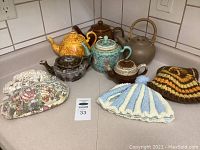 Photo showing five assorted teapots and three tea cozies on a countertop.